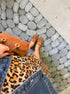 A top-down lifestyle shot of a person wearing a leopard print skirt, blue denim jacket, and brown suede fringed shoes, holding a brown suede handbag on a stone paved path.