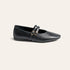 A single black leather flat shoe with a pointed toe and double strap embellishment with gold-tone buckles, shown from a side profile on a light beige background.