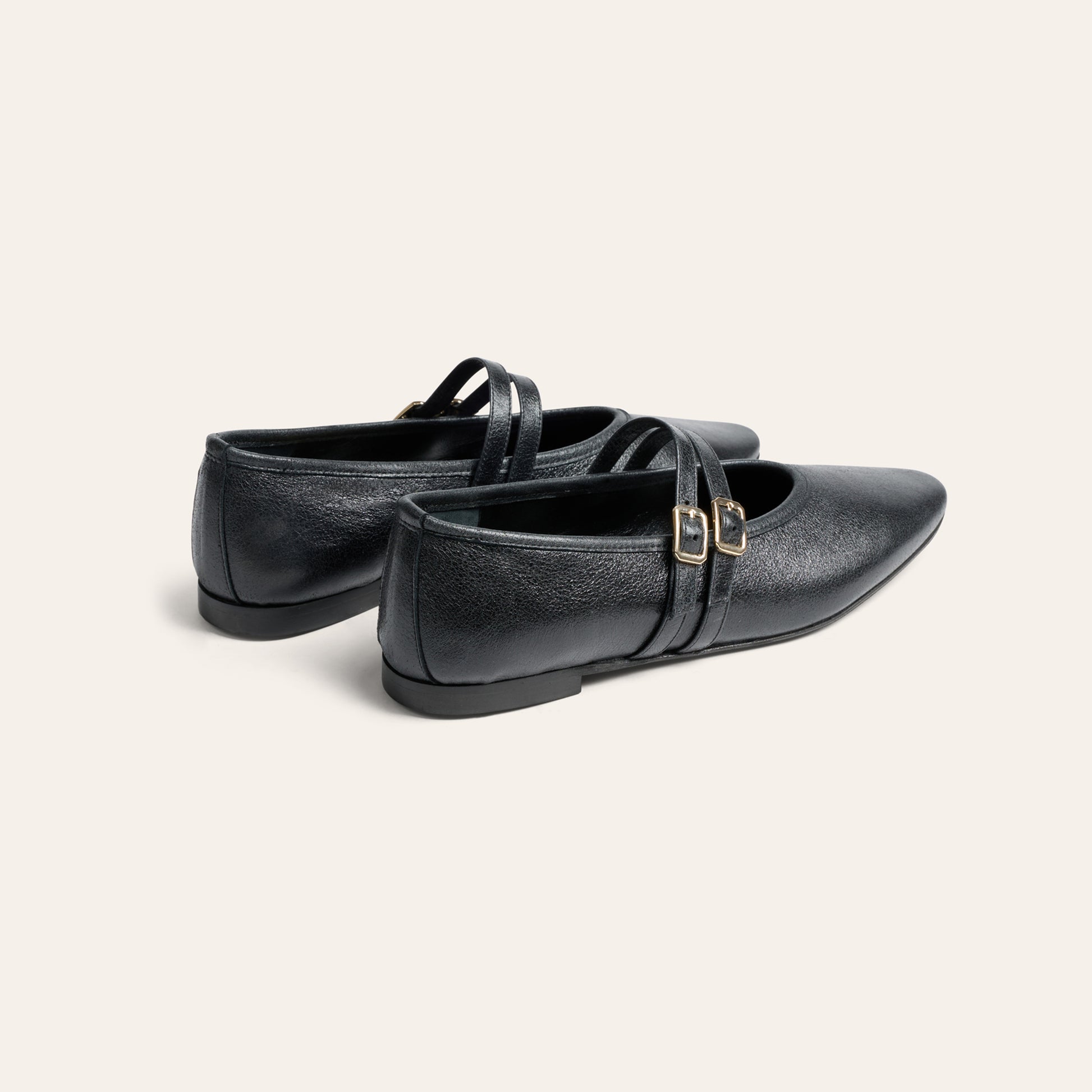 A pair of black leather Mary Jane flats with double straps and small gold-tone buckle details, shown from a three-quarters angle on a light cream background.