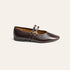 A dark brown leather flat shoe with double straps and gold-tone buckle details, shown from a side profile on a light cream background.