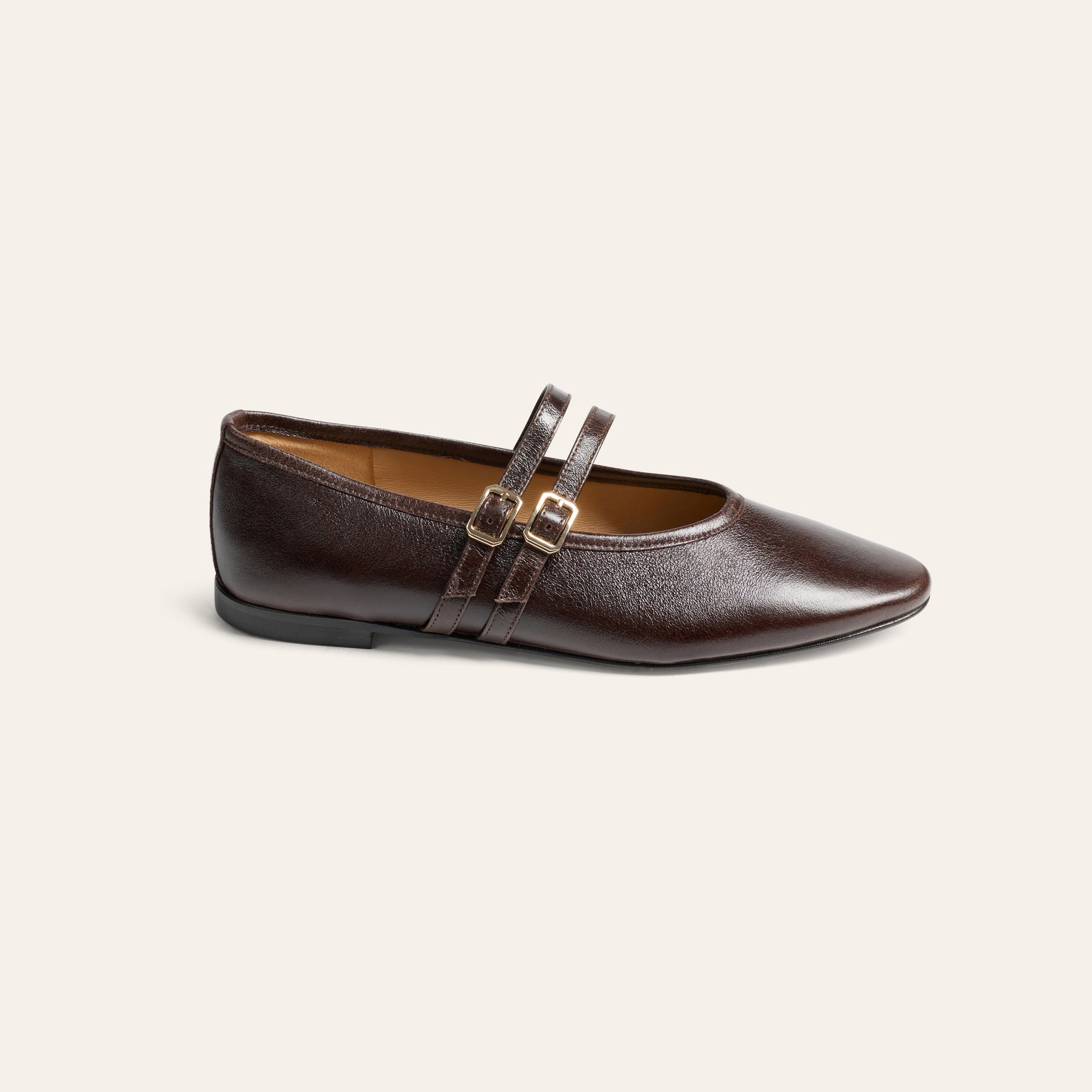 A dark brown leather flat shoe with double straps and gold-tone buckle details, shown from a side profile on a light cream background.