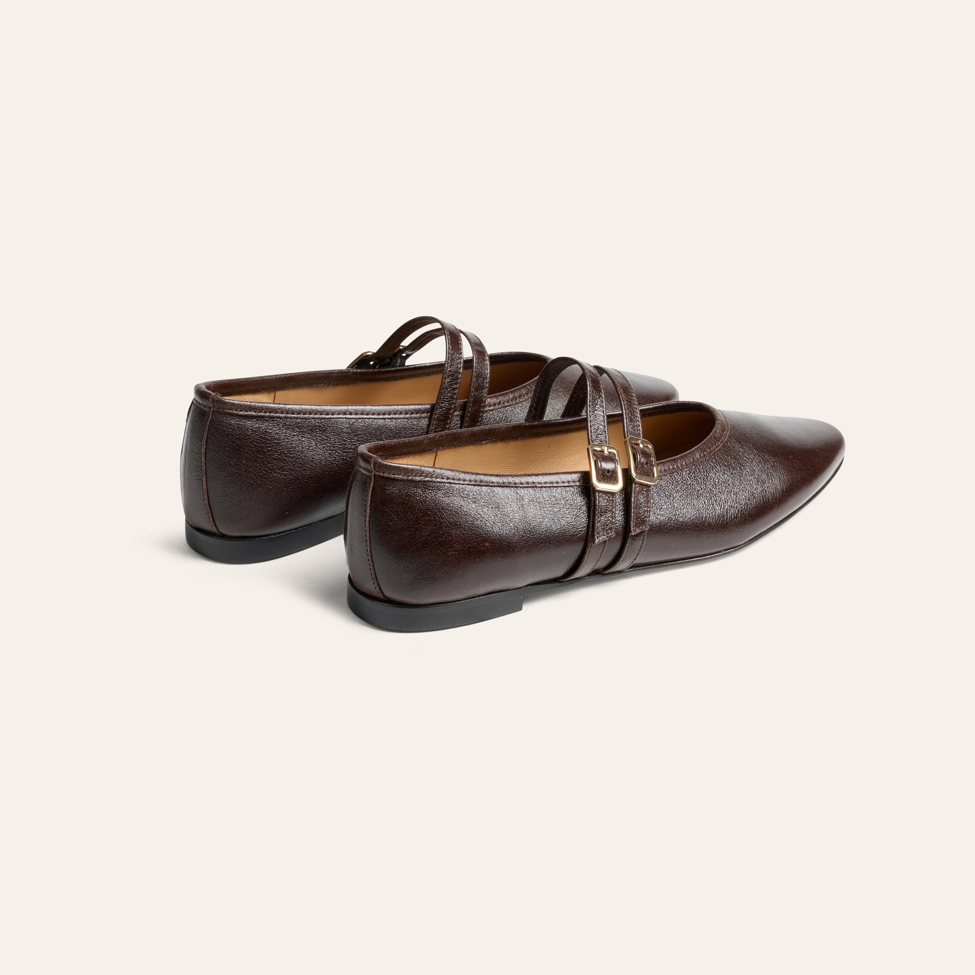 A pair of dark brown leather Mary Jane flats with double straps and gold-tone buckle details, shown on a light cream background.