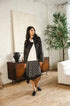 A woman stands in a bright, modern room wearing a black leather jacket, white blouse, black-and-white plaid skirt, and black loafers, holding a black handbag.