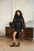 A woman stands in a room wearing a long brown trench coat over a black turtleneck dress, styled with a black belt and dark brown ankle boots. She is posed in front of a wooden sideboard against a textured beige wall.