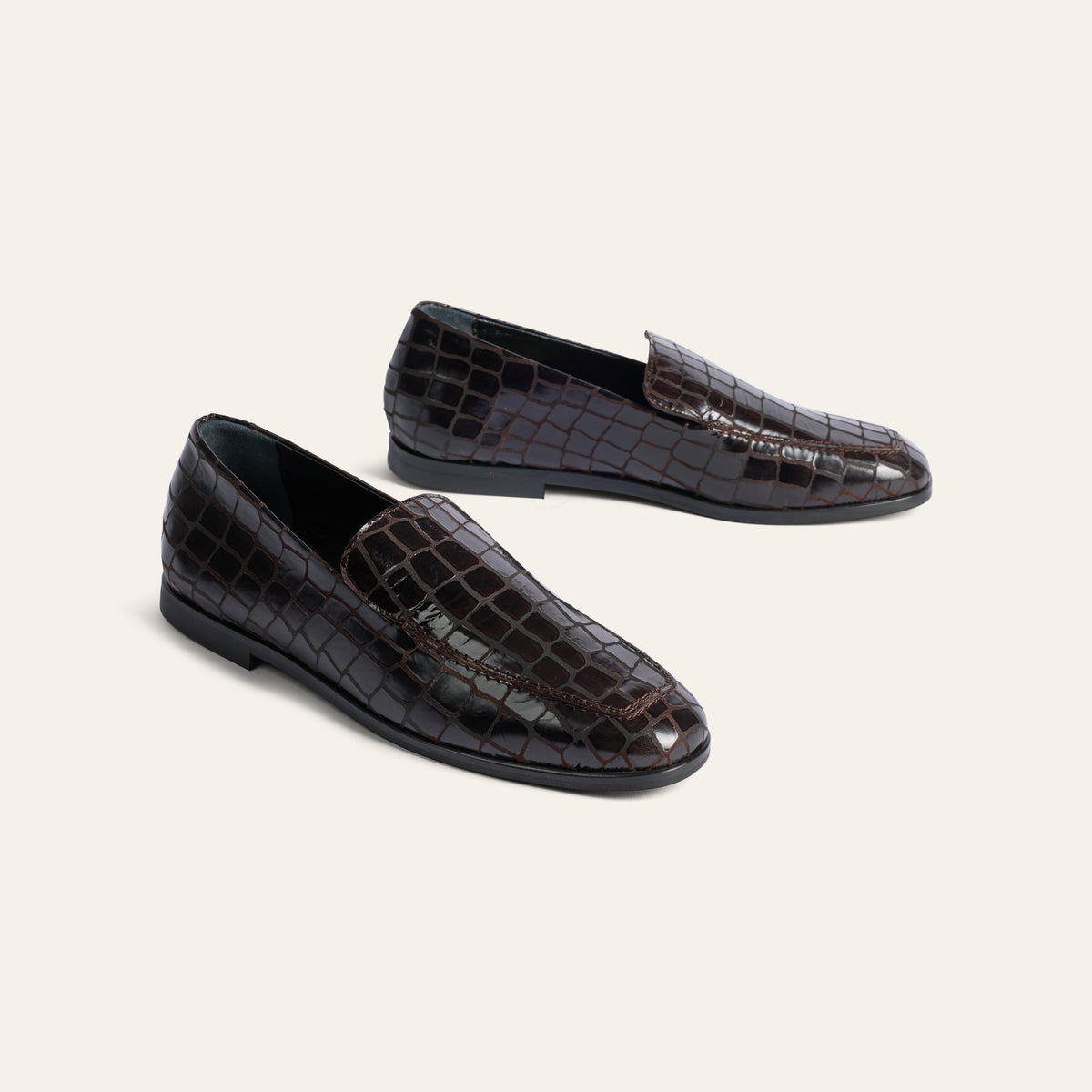 A pair of dark brown crocodile-embossed leather loafers on a light beige background.