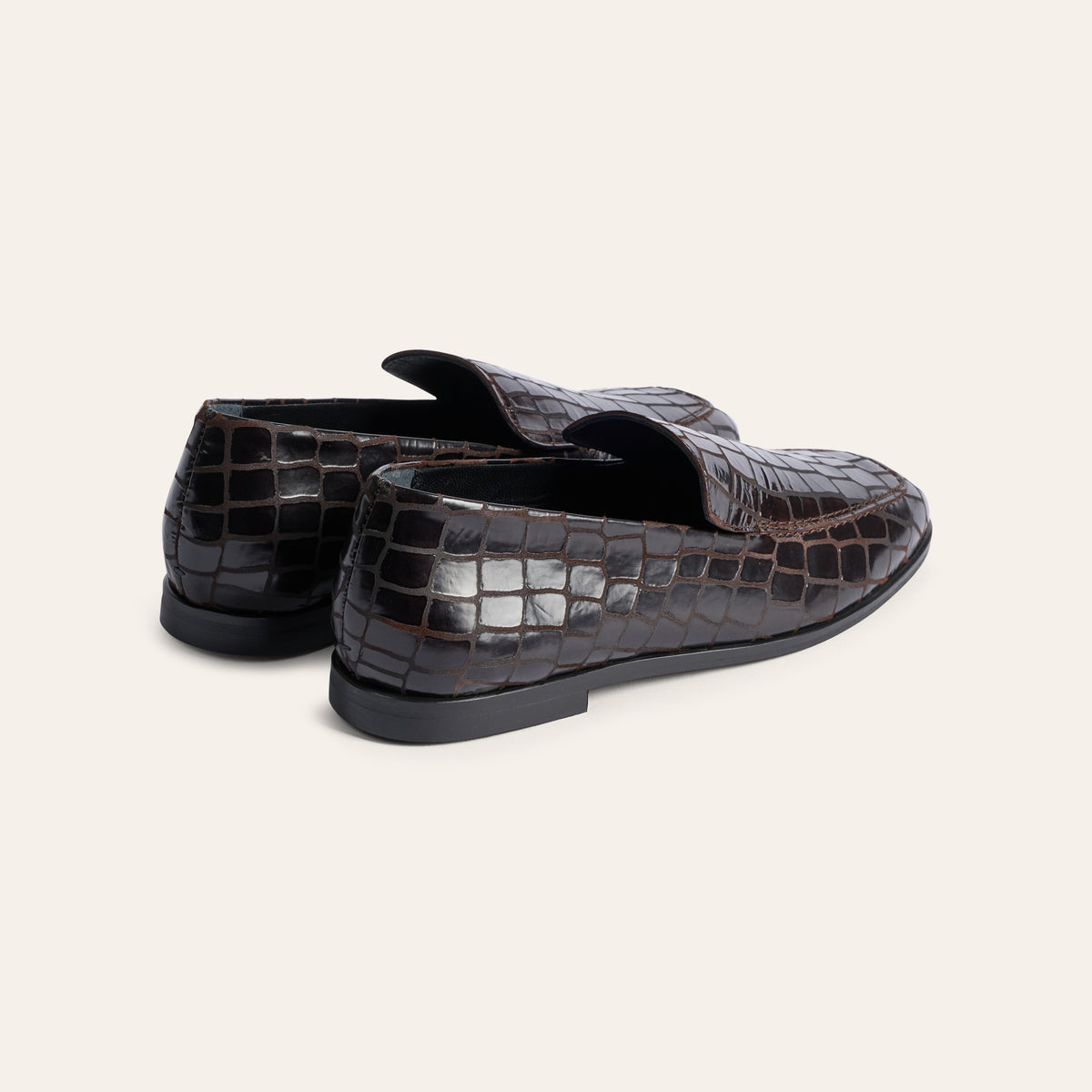 A pair of dark brown crocodile-embossed leather loafers shown from a three-quarters rear angle on a light beige background.