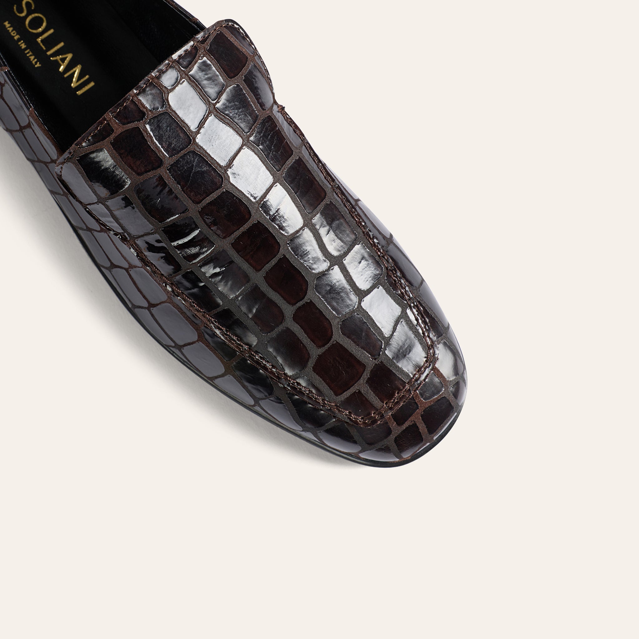 A close-up of a dark brown crocodile-embossed leather loafer by Soliani, showing the glossy textured scales and contrast stitching against a light beige background.