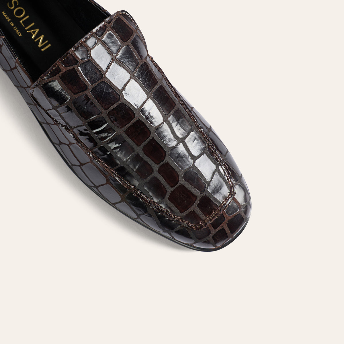 A close-up of a dark brown crocodile-embossed leather loafer by Soliani, showing the glossy textured scales and contrast stitching against a light beige background.