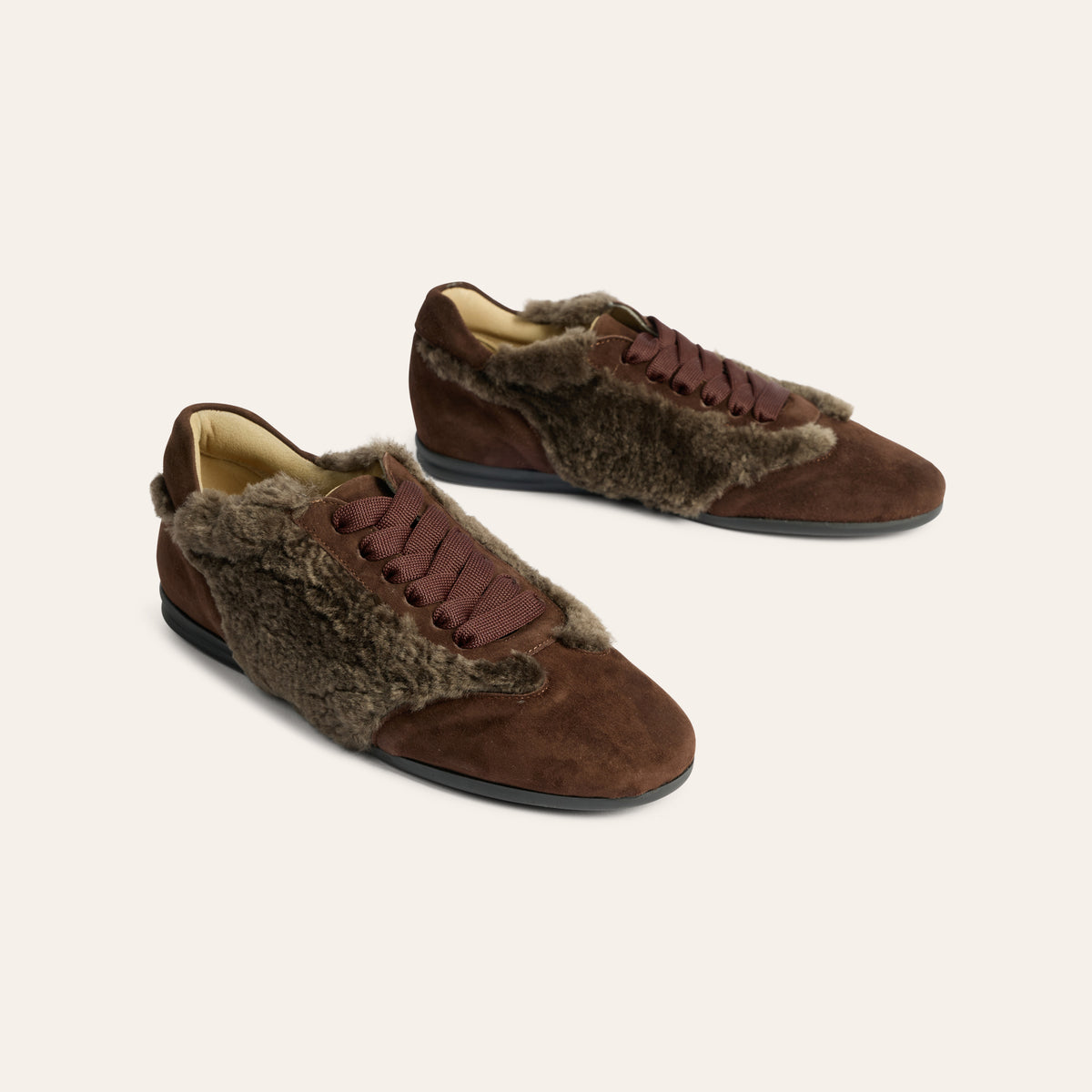 A pair of brown suede slip-on shoes with brown faux-fur trim and matching laces, displayed on a light beige background.