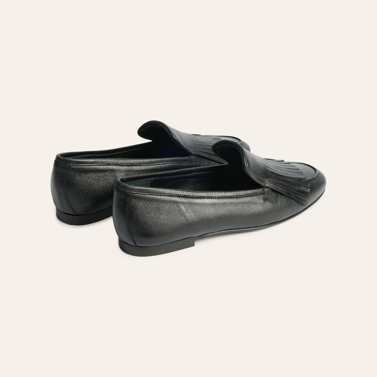 A pair of black leather penny loafers with a tassel detail, shown from a three-quarters rear angle on a light gray studio background.