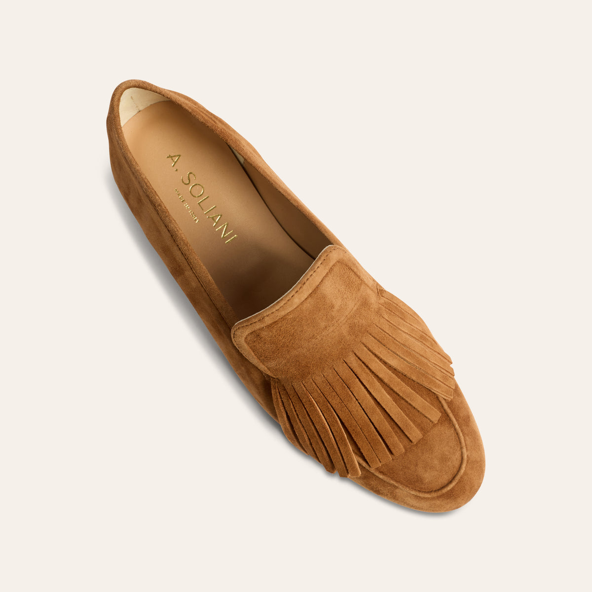 A single tan suede moccasin loafer with fringe detailing, shown from a top-down perspective on a light cream background. The brand name 'A.