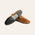 A pair of tan suede fringed loafers shown on a light cream background, with one shoe standing upright to display the black sole and the other lying flat to show the side profile.