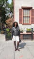 A woman stands on a city sidewalk wearing a black leather jacket, white pleated skirt, and brown over-the-knee boots, with a house and fence in the background.