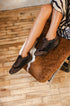 A person seated on a chair wearing dark brown suede and faux-fur lace-up shoes, with a wood plank floor and a brown-and-white animal hide chair visible.