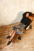 A woman reclines on a modern leather lounge chair in a minimalist room with a textured beige wall and light wood plank floor. She is wearing a black long-sleeve top, a black and beige zebra-print midi skirt, and black slip-on shoes.