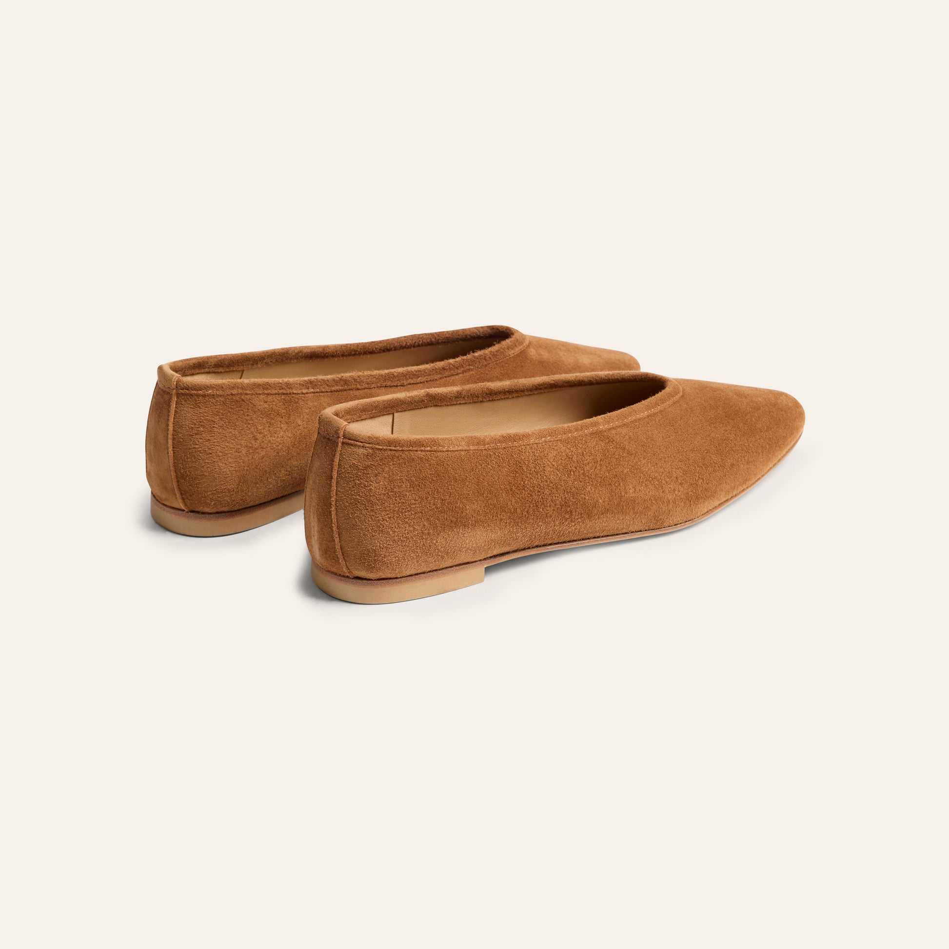 A pair of tan suede flat shoes with pointed toes, shown from a three-quarters angle on a light cream background.