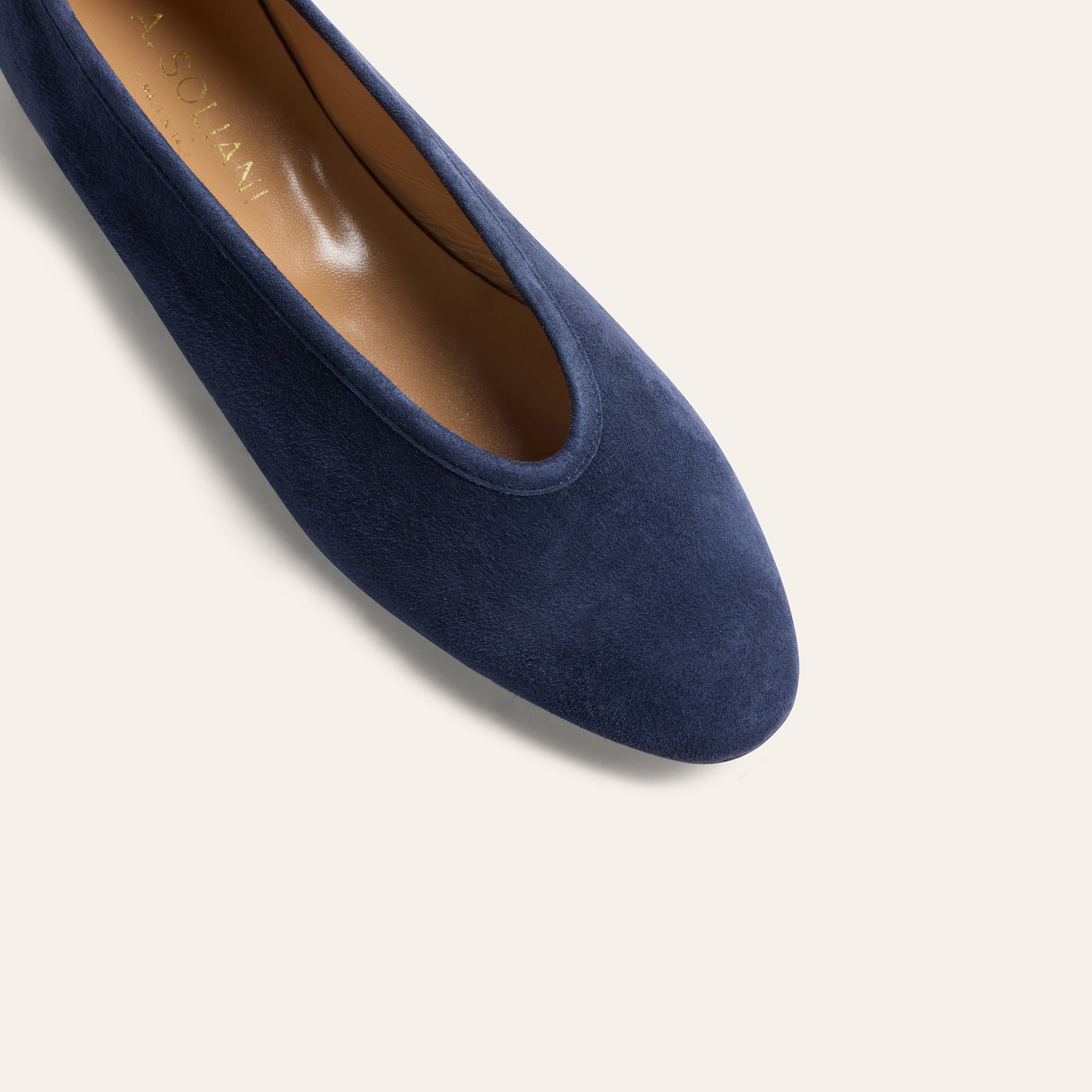A close-up of a navy blue suede flat shoe, showing the textured suede upper, rounded toe, and tan insole with the brand name printed in gold.