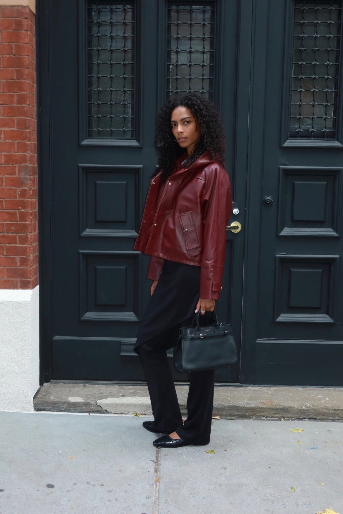 A woman stands on a sidewalk in front of dark green double doors, wearing a cropped burgundy leather jacket, black pants, and holding a black handbag.