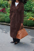 A person wearing a long brown suede-like double-breasted coat over a white top and black pants, holding a tan leather handbag outdoors on a paved walkway with greenery in the background.