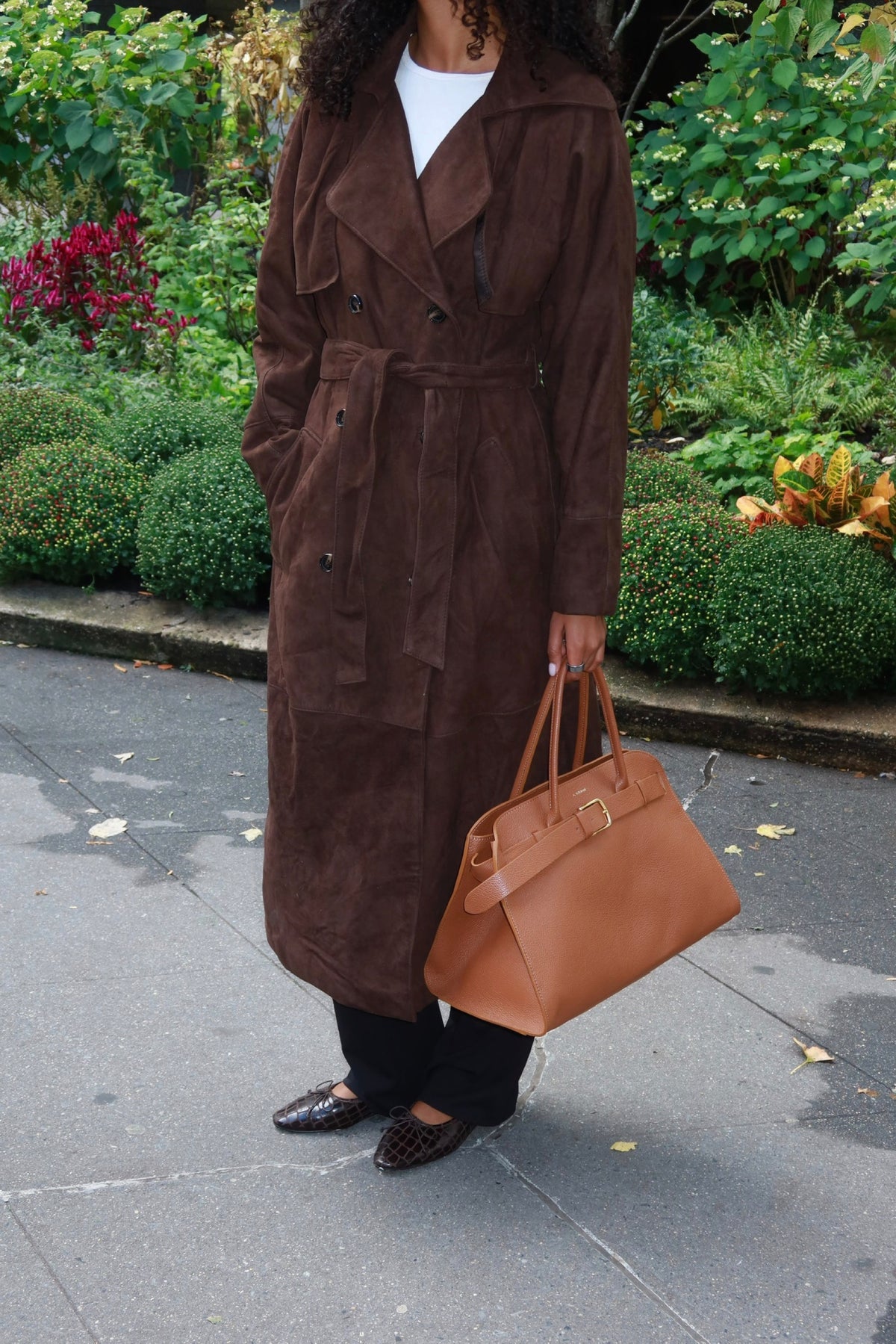 A person wearing a long brown suede-like double-breasted coat over a white top and black pants, holding a tan leather handbag outdoors on a paved walkway with greenery in the background.