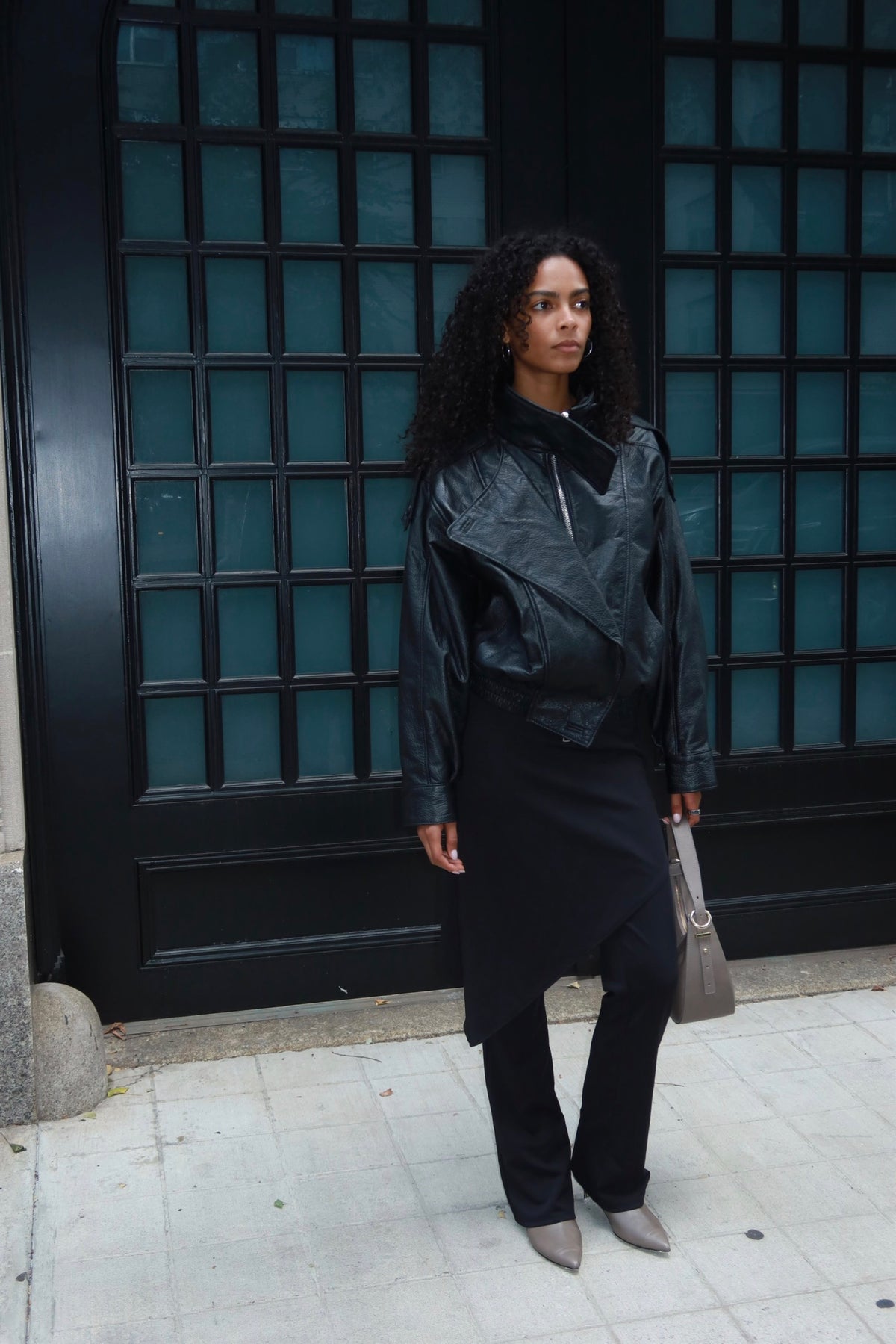 A woman stands on a sidewalk in front of a large black door with blue-tinted glass panels, wearing a black leather jacket, black skirt, black pants, and gray pointed shoes, while carrying a taupe handbag.