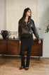 A woman stands in a room wearing a dark brown leather jacket, a black asymmetrical skirt, black trousers, and red shoes. She is posed in front of a wooden cabinet and textured wall.
